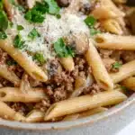 One pot pasta mit hack und pilzen - the image is a close-up of a bowl of penne pasta. the pasta is cooked al dente and is topped with a generous amount of ground beef, sliced mushrooms, and a sprinkle of parmesan cheese. the dish is garnished with fresh parsley. the bowl is made of ceramic and is sitting on a white surface. the background is blurred, making the pasta the focal point of the image.