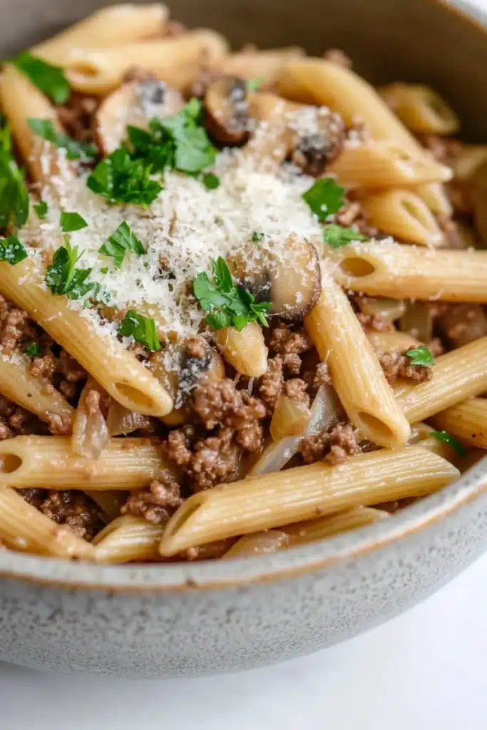 One pot pasta mit hack und pilzen - the image is a close-up of a bowl of penne pasta. the pasta is cooked al dente and is topped with a generous amount of ground beef, sliced mushrooms, and a sprinkle of parmesan cheese. the dish is garnished with fresh parsley. the bowl is made of ceramic and is sitting on a white surface. the background is blurred, making the pasta the focal point of the image.