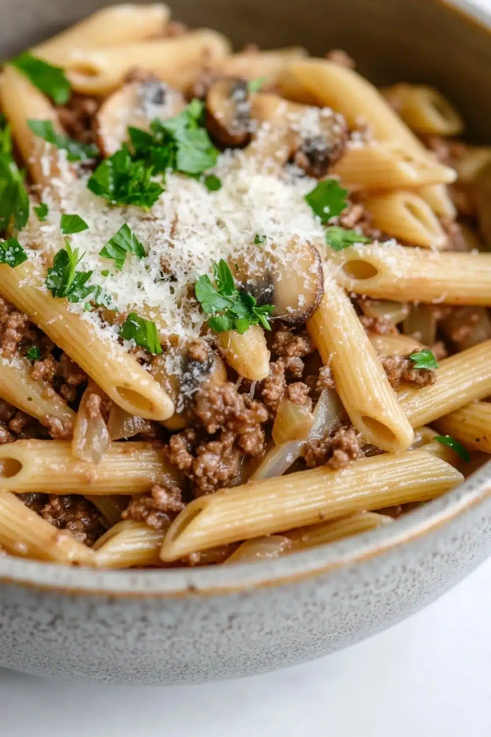One pot pasta mit hack und pilzen - the image is a close-up of a bowl of penne pasta. the pasta is cooked al dente and is topped with a generous amount of ground beef, sliced mushrooms, and a sprinkle of parmesan cheese. the dish is garnished with fresh parsley. the bowl is made of ceramic and is sitting on a white surface. the background is blurred, making the pasta the focal point of the image.