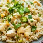 One pot queso chicken rice - the image is a close-up of a plate of rice dish. the rice appears to be cooked and has a light brown color. on top of the rice, there are chunks of chicken, green peas, and yellow bell peppers. the dish is garnished with fresh cilantro leaves. the plate is sitting on a wooden table.
