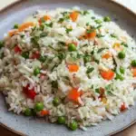 One pot rice - the image is a close-up of a plate of rice dish. the plate is round and has a light brown color with a speckled texture. the rice is white and fluffy, and it is mixed with various vegetables such as peas, carrots, and red bell peppers. the vegetables are garnished with chopped parsley. the dish appears to be freshly cooked and ready to eat.
