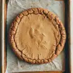 Peanut butter cookie crust dessert - the image shows a freshly baked pie in a rectangular baking tray. the pie has a golden brown crust on top and appears to be freshly baked. the crust is crinkled and has a crumbly texture. the tray is lined with parchment paper, which is resting on a white wooden surface. the background is blurred, but it seems to be a kitchen countertop.