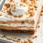 Peanut butter cookie layered dessert - the image is of a slice of dessert on a white rectangular baking tray. the dessert appears to be a layered dessert with a layer of whipped cream on top. the cream is a light brown color and is drizzled over the top with a crumbly texture. there are also some nuts scattered around the dessert, adding a crunchy texture. the tray is sitting on a wooden table.