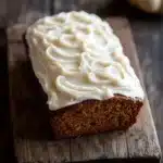 Pumpkin bread icing - the image shows a rectangular loaf of cake on a wooden cutting board. the cake appears to be freshly baked and has a golden brown color. on top of the cake, there is a generous layer of white frosting that is swirled in a swirl pattern. the frosting is smooth and glossy, and the cake looks moist and fluffy. in the background, there are two small white pumpkins, one on the right side of the image and the other on the left side. the cutting board is placed on a dark wooden table.