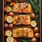 Sheet pan lemon brown butter salmon - the image shows a rectangular baking tray with two pieces of salmon on it. the salmon is cooked to a golden brown color and appears to be seasoned with herbs and spices. the tray is filled with various vegetables, including green beans, potatoes, carrots, and lemons. the vegetables are arranged in a colorful and appetizing manner, with some of them overlapping each other. the background is a wooden table, and the tray is lined with parchment paper.