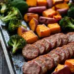 Sheet pan sausage sweet potato broccoli - the image shows a baking tray filled with various types of food. on the left side of the tray, there are roasted sweet potatoes, broccoli, and sausage. the sweet potatoes are orange in color and have a crispy texture. the broccoli is bright green and looks fresh. the sausage is cut into thin slices and is arranged in a single layer on the tray. the tray is lined with parchment paper and is sitting on a wooden table.