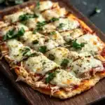Shredded chicken keto pizza crust - the image shows a freshly baked pizza on a wooden cutting board. the pizza has a golden brown crust and is topped with melted cheese and fresh herbs. the cheese is melted and bubbly, and the herbs are scattered around the edges of the pizza. there are also a few cherry tomatoes and a bunch of arugula on the cutting board, as well as a few garlic cloves. the background is a dark grey countertop.