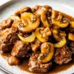 Slow cooker beef mushroom - the image shows a plate of food with a dish of beef and mushrooms. the beef appears to be cooked medium-rare and is covered in a rich, dark brown sauce. the mushrooms are sliced and arranged in a pile on top of the beef. the sauce is a light brown color and is drizzled over the top. the plate is white with a speckled pattern around the edges. the background is blurred, but it seems to be a kitchen countertop.