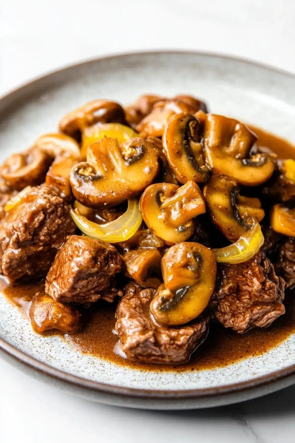 Slow cooker beef mushroom - the image shows a plate of food with a dish of beef and mushrooms. the beef appears to be cooked medium-rare and is covered in a rich, dark brown sauce. the mushrooms are sliced and arranged in a pile on top of the beef. the sauce is a light brown color and is drizzled over the top. the plate is white with a speckled pattern around the edges. the background is blurred, but it seems to be a kitchen countertop.