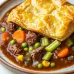 Slow cooker beef pot pie - the image shows a plate of beef stew with a golden brown crust on top. the stew is made with chunks of beef, peas, carrots, celery, and corn. the beef is cooked to a medium-rare and appears to be tender and juicy. the peas are bright green and the carrots are orange. the corn is yellow and is scattered throughout the stew. the plate is brown and has a speckled pattern around the edges. the background is a white marble countertop.