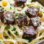 Steak bites with garlic parmesan pasta - the image is a close-up of a bowl of pasta dish. the pasta appears to be fettuccine alfredo, which is a type of italian pasta. the dish is served in a brown ceramic bowl and is garnished with chopped parsley. on top of the pasta, there are several pieces of cooked meat, which appear to be beef or beef tenderloin. the meat is covered in a creamy white sauce, which looks like it has been cooked to perfection. the sauce is drizzled over the top, giving the dish a glossy sheen. the bowl is sitting on a wooden table, and the background is blurred, making the dish the focal point of the image.
