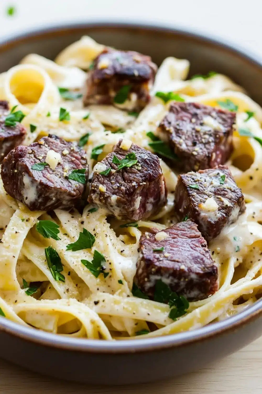 Steak bites with garlic parmesan pasta - the image is a close-up of a bowl of pasta dish. the pasta appears to be fettuccine alfredo, which is a type of italian pasta. the dish is served in a brown ceramic bowl and is garnished with chopped parsley. on top of the pasta, there are several pieces of cooked meat, which appear to be beef or beef tenderloin. the meat is covered in a creamy white sauce, which looks like it has been cooked to perfection. the sauce is drizzled over the top, giving the dish a glossy sheen. the bowl is sitting on a wooden table, and the background is blurred, making the dish the focal point of the image.