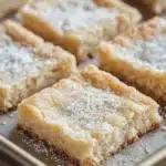 Sugar cookie bar - the image shows a tray of freshly baked square bars. the bars are golden brown in color and have a crumbly texture. they are dusted with powdered sugar, giving them a shiny appearance. the tray is lined with parchment paper and is placed on a wooden table. the background is blurred, but it appears to be a kitchen countertop.