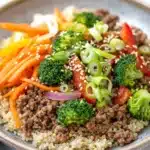 Viral high protein beef bowl - the image is a close-up of a plate of food. the plate is made of a light-colored ceramic bowl with a gray rim. the food appears to be a stir-fry dish with a variety of ingredients, including sliced carrots, broccoli, red bell peppers, and sliced green onions. the vegetables are arranged in a colorful and appetizing manner, with some overlapping each other. the ground beef is a dark brown color, and the vegetables are a mix of different shades of green, red, and yellow. the dish is garnished with sesame seeds, giving it a crunchy texture. the background is white, making the colors of the food stand out.