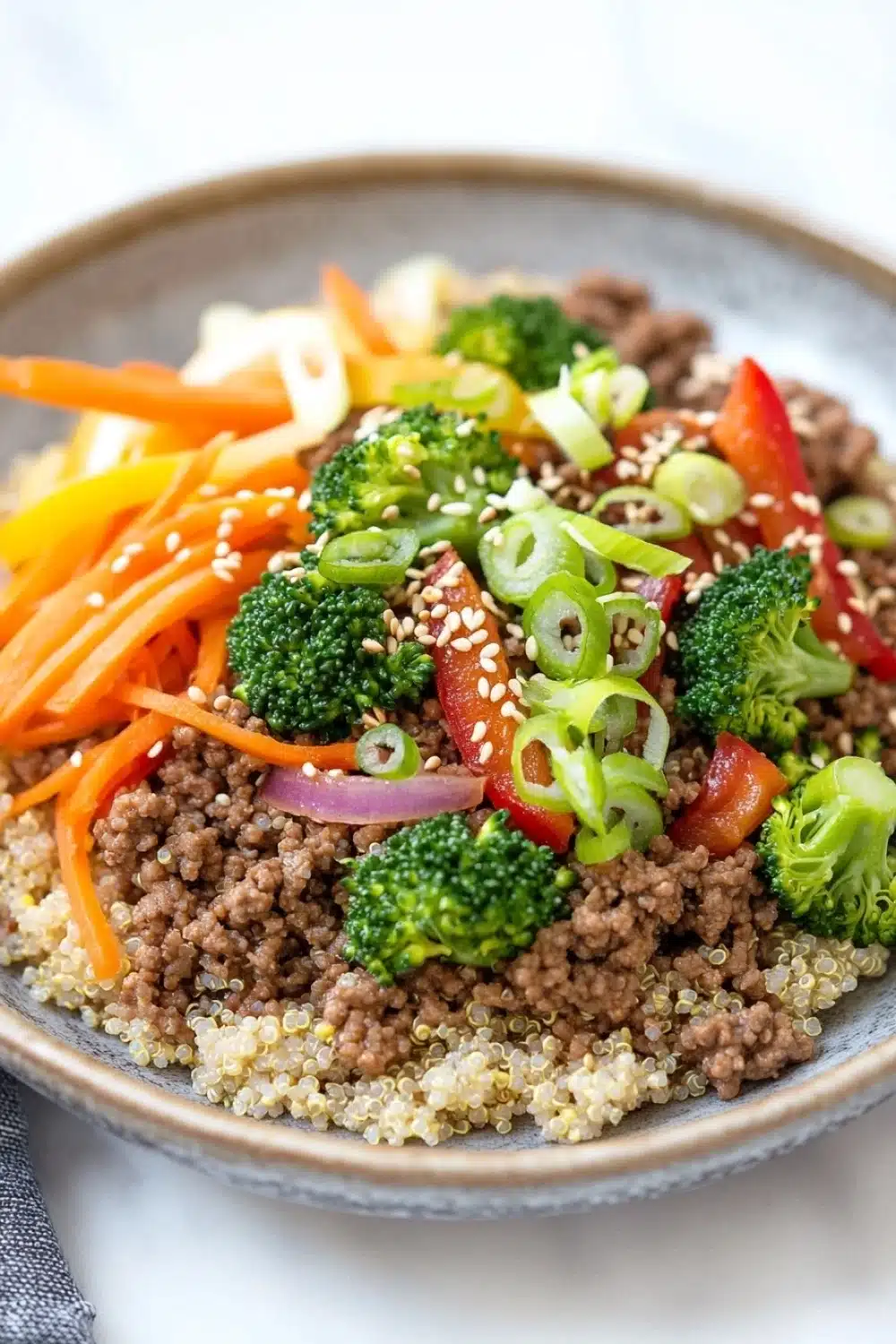 Viral high protein beef bowl - the image is a close-up of a plate of food. the plate is made of a light-colored ceramic bowl with a gray rim. the food appears to be a stir-fry dish with a variety of ingredients, including sliced carrots, broccoli, red bell peppers, and sliced green onions. the vegetables are arranged in a colorful and appetizing manner, with some overlapping each other. the ground beef is a dark brown color, and the vegetables are a mix of different shades of green, red, and yellow. the dish is garnished with sesame seeds, giving it a crunchy texture. the background is white, making the colors of the food stand out.