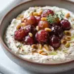 Whipped feta dip with roasted grapes - the image is a close-up of a bowl of hummus. the bowl is made of ceramic and is placed on a white marble countertop. the hummus is creamy and has a light brown color. on top of the hummus, there are several red cherries arranged in a circular pattern. the cherries are garnished with a sprig of rosemary and pine nuts. there are also a few scattered pine nuts on the countertop next to the bowl. the overall presentation of the dish is appetizing and visually appealing.