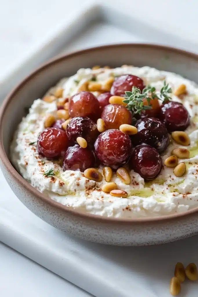 Whipped feta dip with roasted grapes - the image is a close-up of a bowl of hummus. the bowl is made of ceramic and is placed on a white marble countertop. the hummus is creamy and has a light brown color. on top of the hummus, there are several red cherries arranged in a circular pattern. the cherries are garnished with a sprig of rosemary and pine nuts. there are also a few scattered pine nuts on the countertop next to the bowl. the overall presentation of the dish is appetizing and visually appealing.