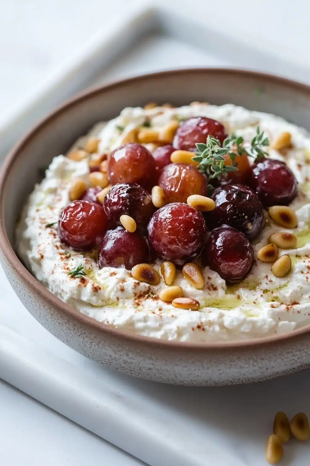 Whipped feta dip with roasted grapes - the image is a close-up of a bowl of hummus. the bowl is made of ceramic and is placed on a white marble countertop. the hummus is creamy and has a light brown color. on top of the hummus, there are several red cherries arranged in a circular pattern. the cherries are garnished with a sprig of rosemary and pine nuts. there are also a few scattered pine nuts on the countertop next to the bowl. the overall presentation of the dish is appetizing and visually appealing.