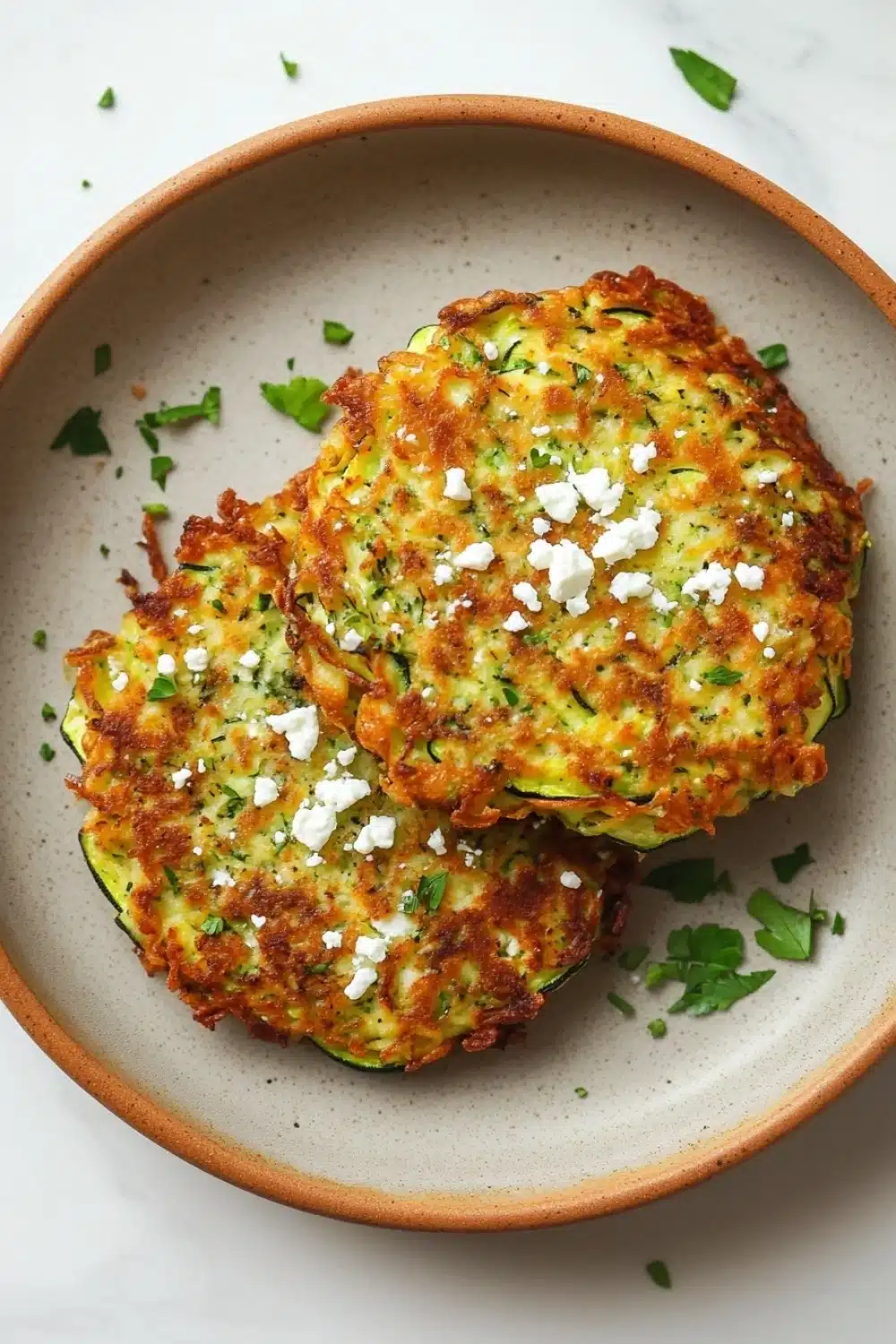 Zucchini feta fritters - the image shows a plate of zucchini fritters on a white marble countertop. the plate is made of terracotta and has a light brown color. the zucchinis are golden brown and appear to be freshly cooked. they are topped with crumbled feta cheese and sprinkled with chopped herbs. there are also a few sprigs of parsley scattered around the plate. the overall presentation of the dish is appetizing and inviting.